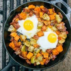 Overhead view of apple and sweet potato hash with two fried eggs in a cast iron skillet over a campfire.
