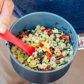 A person in a blue shirt and beige pants is holding a pot of cooked pasta with greens, tomatoes, and yellow squash.