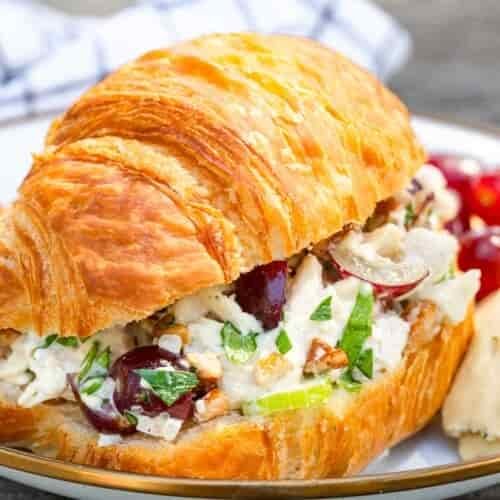 A croissant sandwich with chicken salad, grapes, celery, and pecans on a plate with grapes and a pastry in the background.