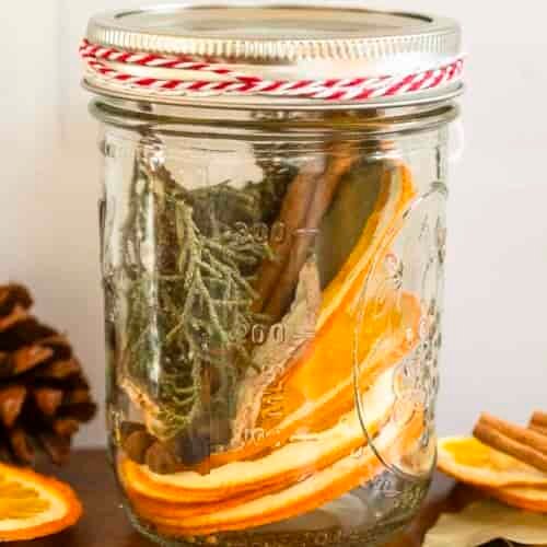 A mason jar with dried orange slices, cinnamon sticks, and greenery sits on wood beside a pinecone and extra dried orange slices.